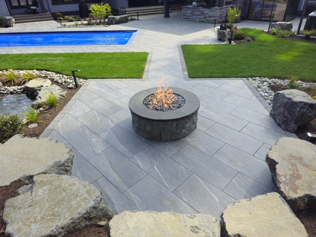 Round stone gas fire pit on a bluestone patio beside a pool in Bellevue at dusk
