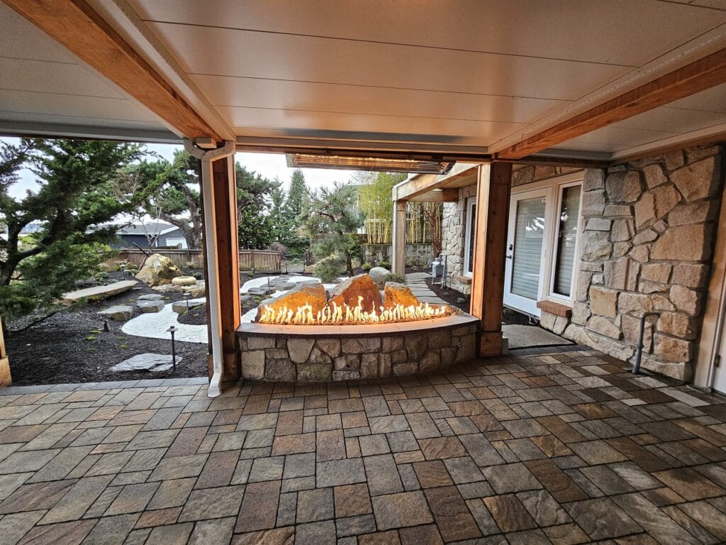 Curved stone gas fireplace with a long ribbon of flame and basalt boulders framed by timber posts facing a garden