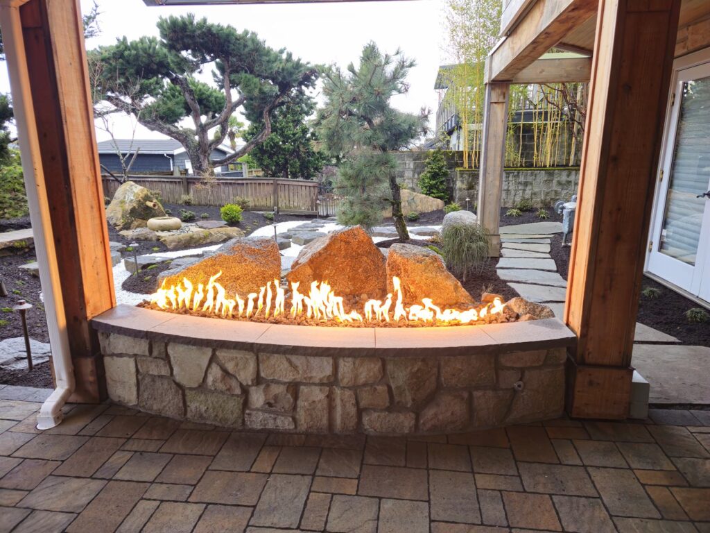 Curved stone gas fireplace with a full ribbon of flame and lava rock viewed across a Japanese-style garden