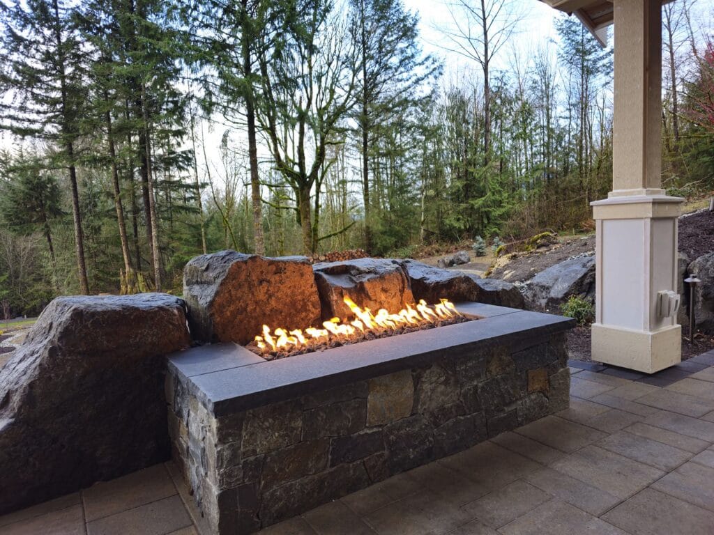 Custom linear gas fire trough built into a stone bench with basalt boulders and forest view