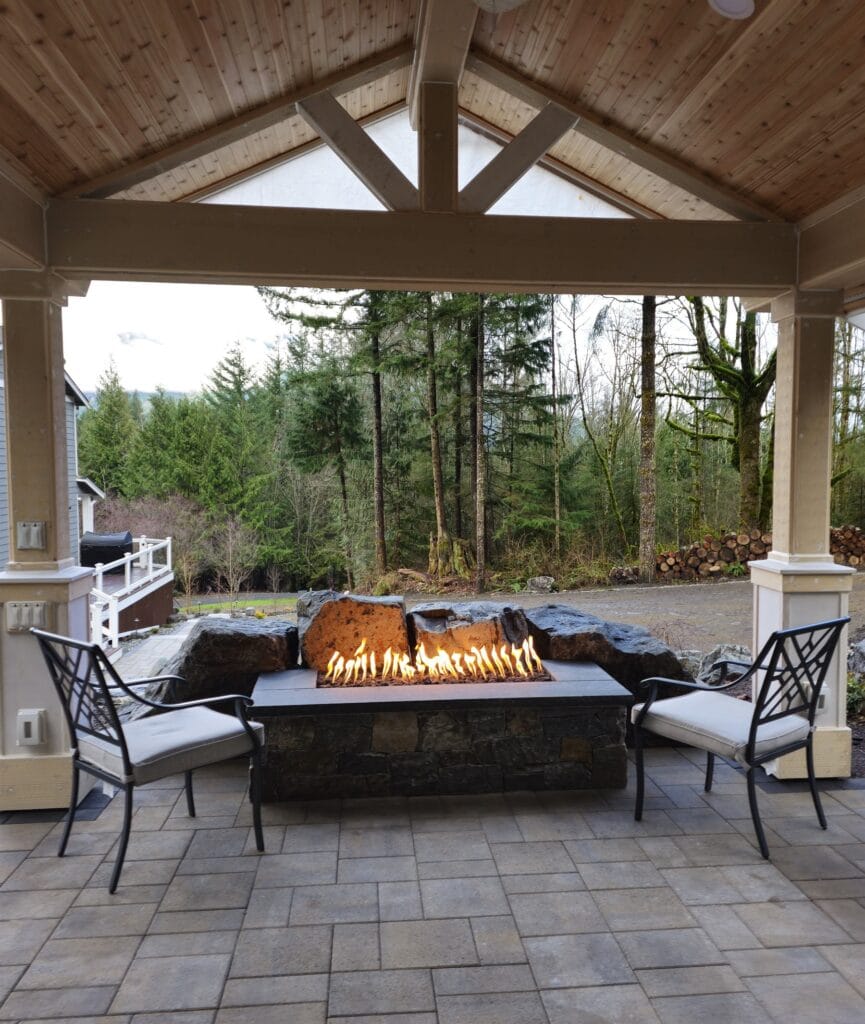 Linear gas fire trough with a low flame framed by two lounge chairs beneath a timber patio cover