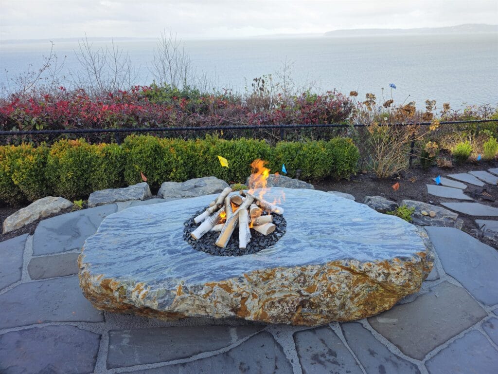 Large flat natural boulder carved into a gas fire pit with a log set on a flagstone patio above the water