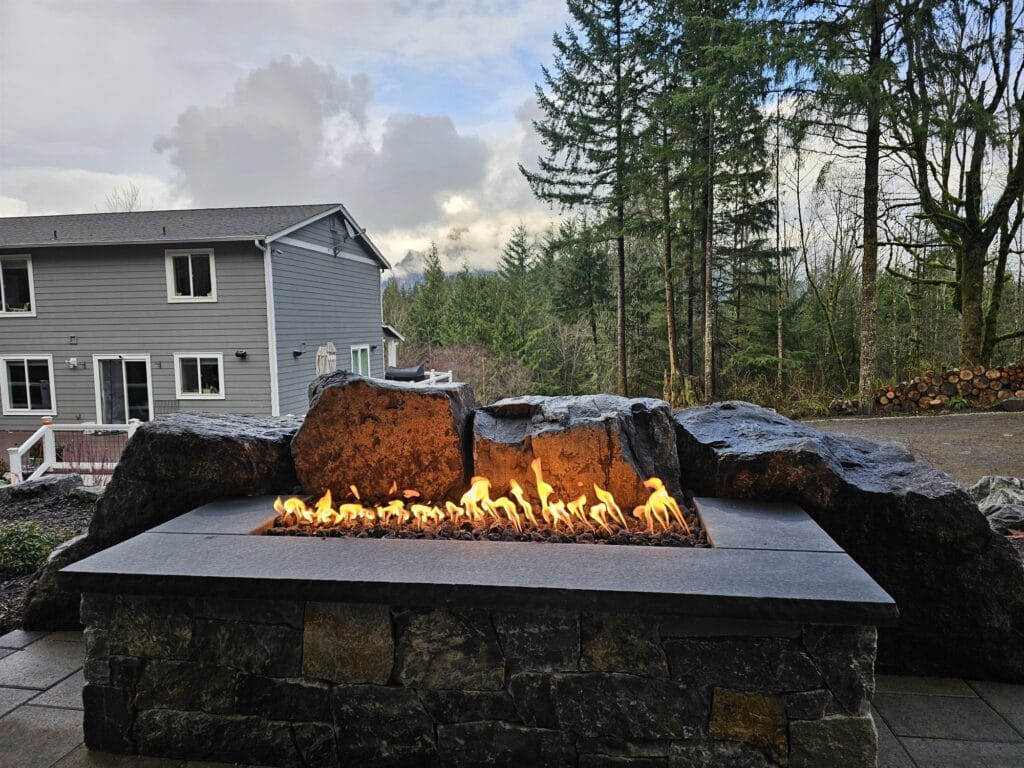 Linear gas fire pit on a stone base with flames rising between large natural boulders