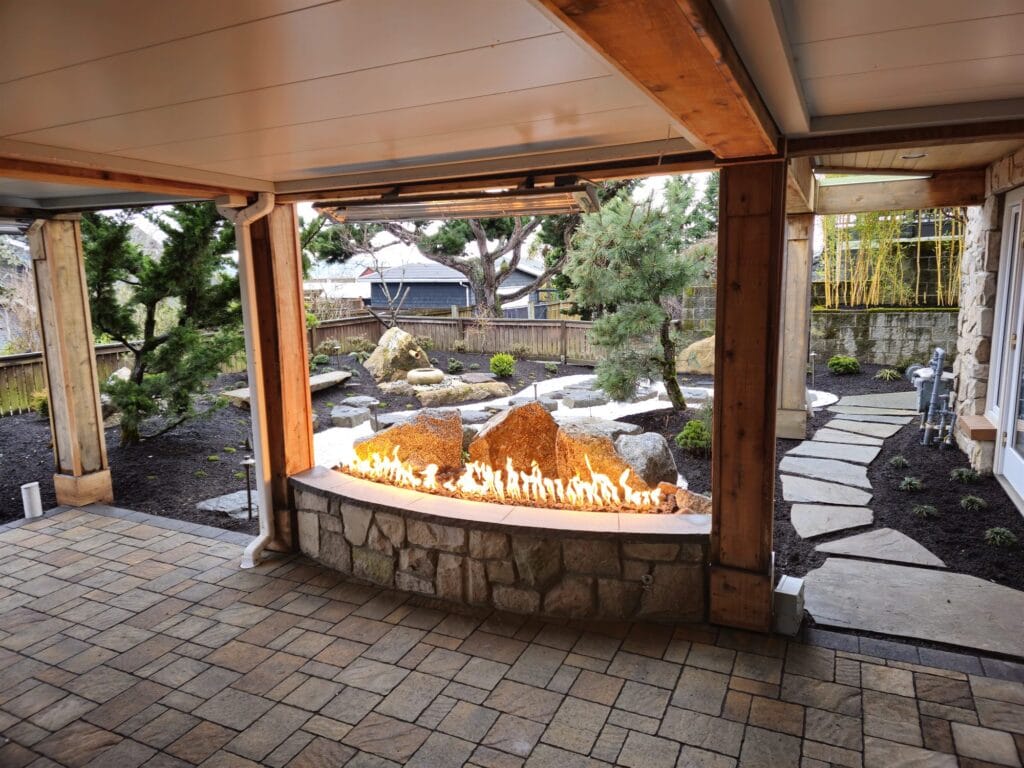 Curved stone gas fire feature with boulders and a ribbon of flame, seen from a covered paver patio