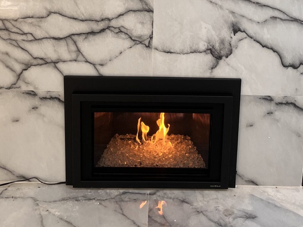 Gas fireplace insert with a black frame and glass media burning, set into a marble-look tile surround