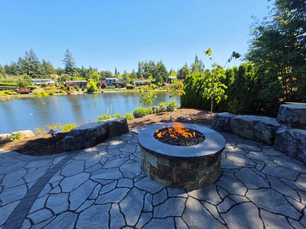 Round gas fire pit on a flagstone patio with boulder seating along a lakeshore
