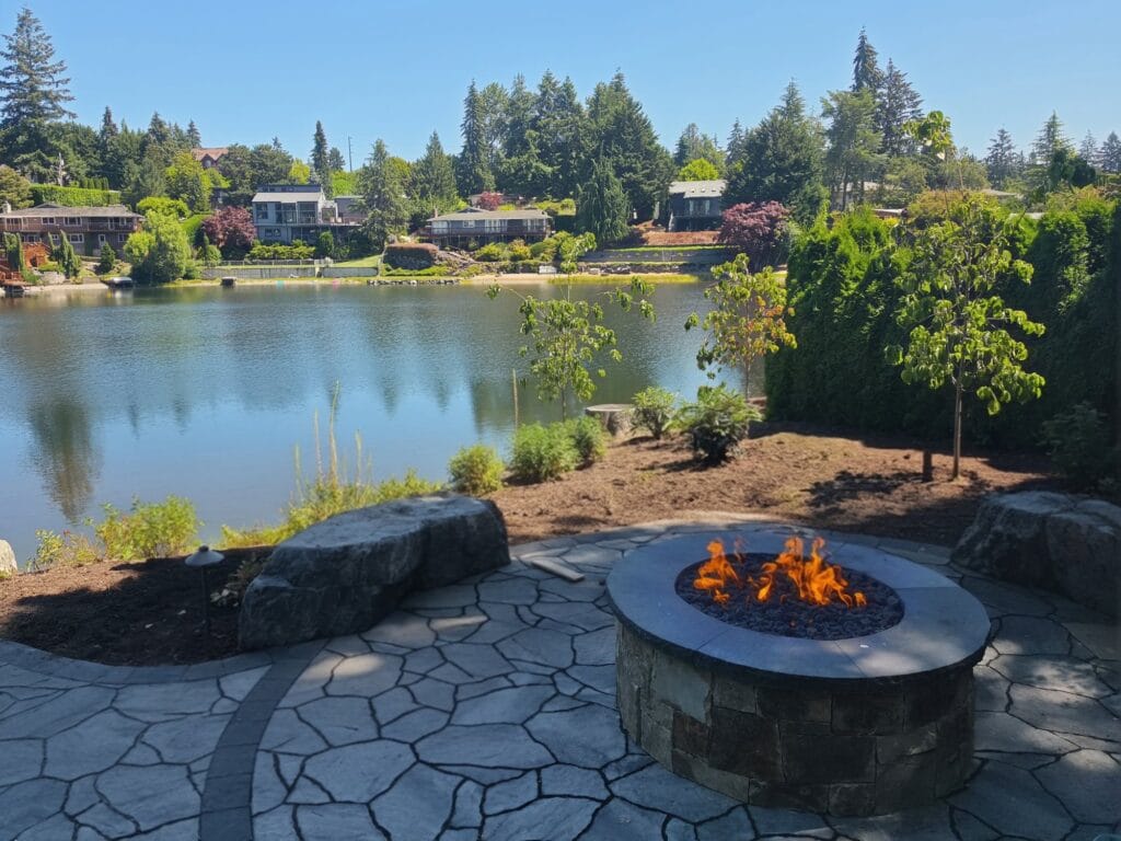 Lakeside flagstone patio with a round stone gas fire pit, boulder seating and new plantings along the shore