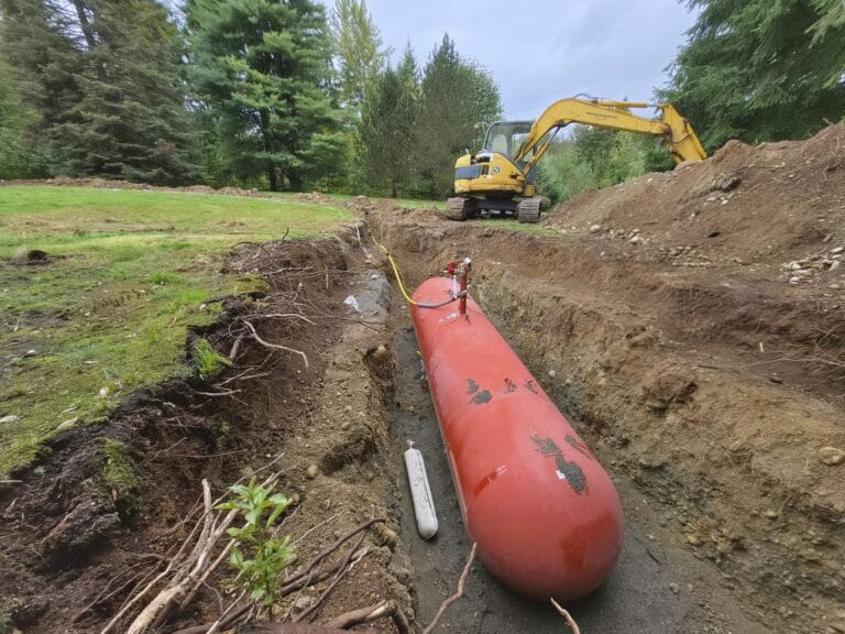 Open trench across a Maple Valley yard with a yellow underground gas line running to a buried propane tank, excavator in the background