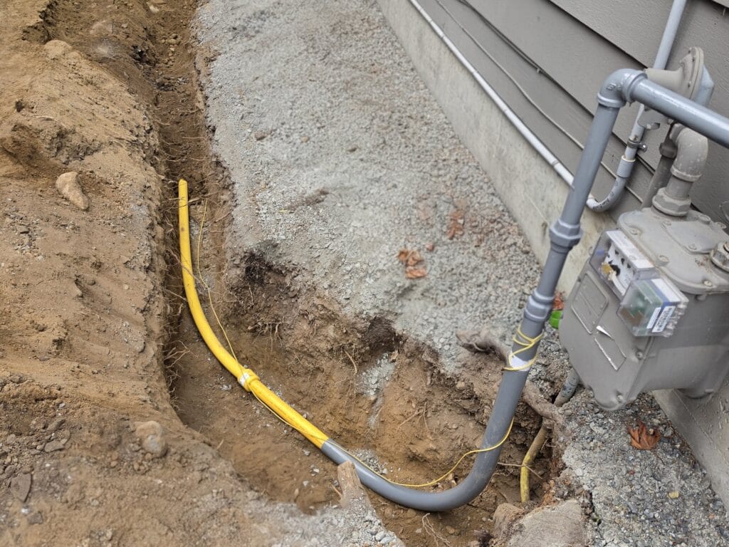 New yellow polyethylene gas line in an open trench, rising to the gas meter on the side of a home