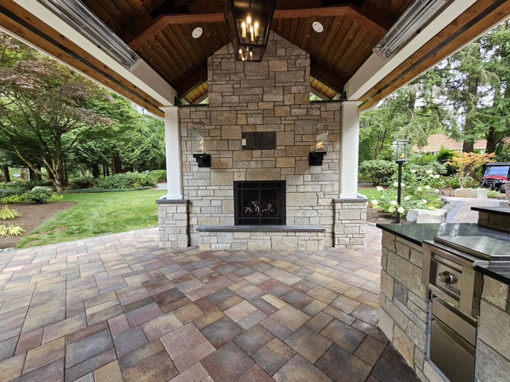 Stone outdoor gas fireplace with wall sconces in a timber pavilion beside a built-in outdoor kitchen