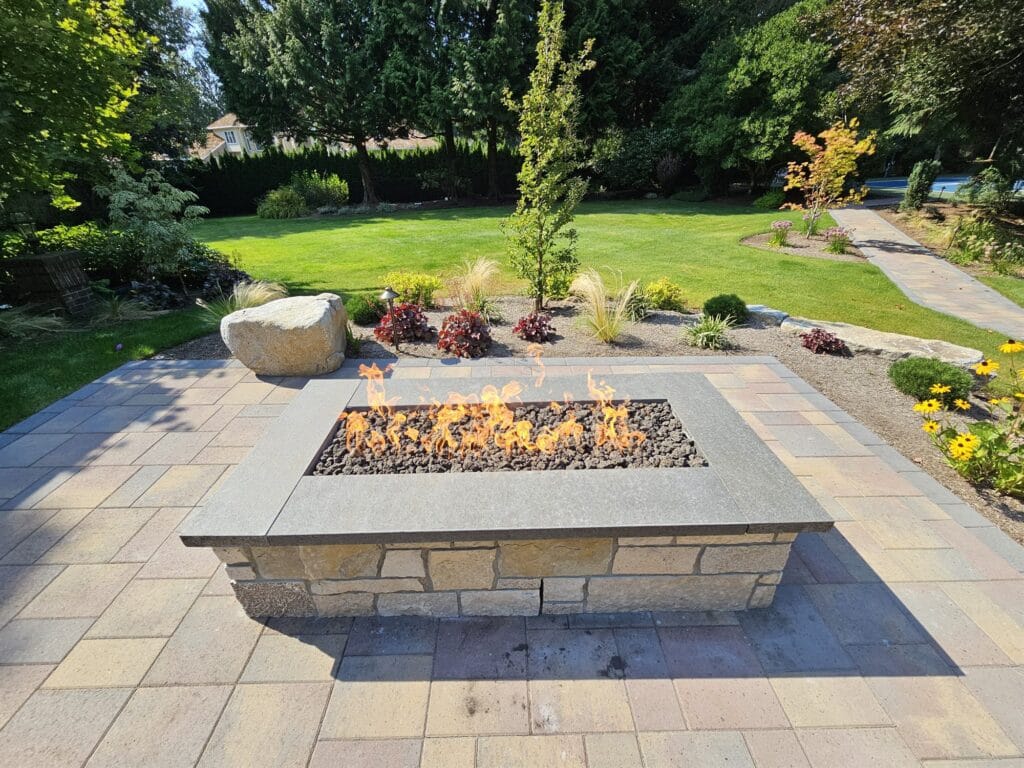 Rectangular stacked-stone gas fire pit with a stone cap and lava rock on a multicolor paver patio