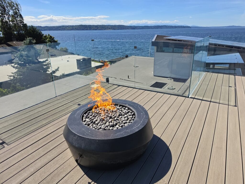 Round dark gas fire bowl filled with lava stones on a rooftop deck with glass railings overlooking the water