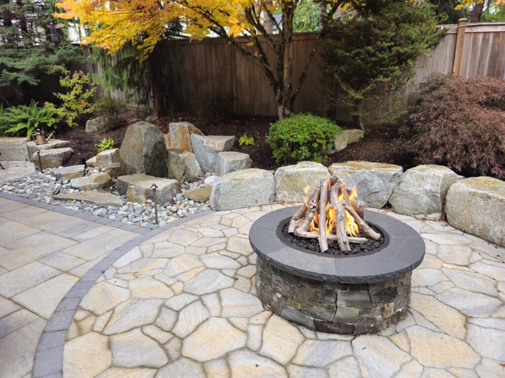 Round stacked-stone gas fire pit with a driftwood log set on a flagstone patio edged with boulders