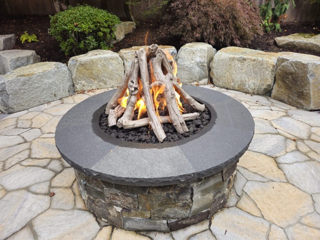 Close view of a driftwood-style gas log set burning in a round stone fire pit with a bluestone cap