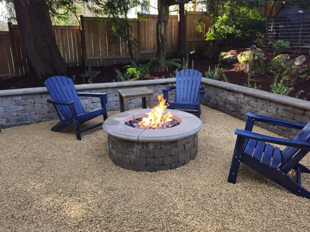 Round block gas fire pit on a gravel patio with blue Adirondack chairs inside a low seat wall