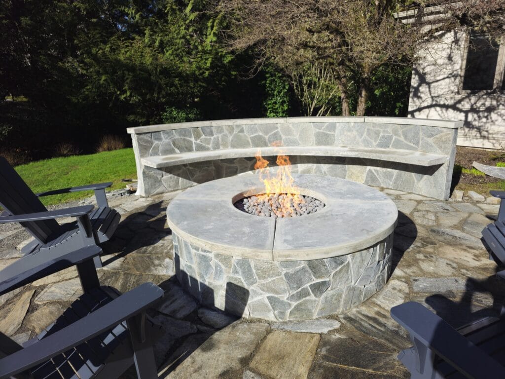 Round flagstone-clad gas fire pit with a wide stone cap in front of a curved stone seat wall