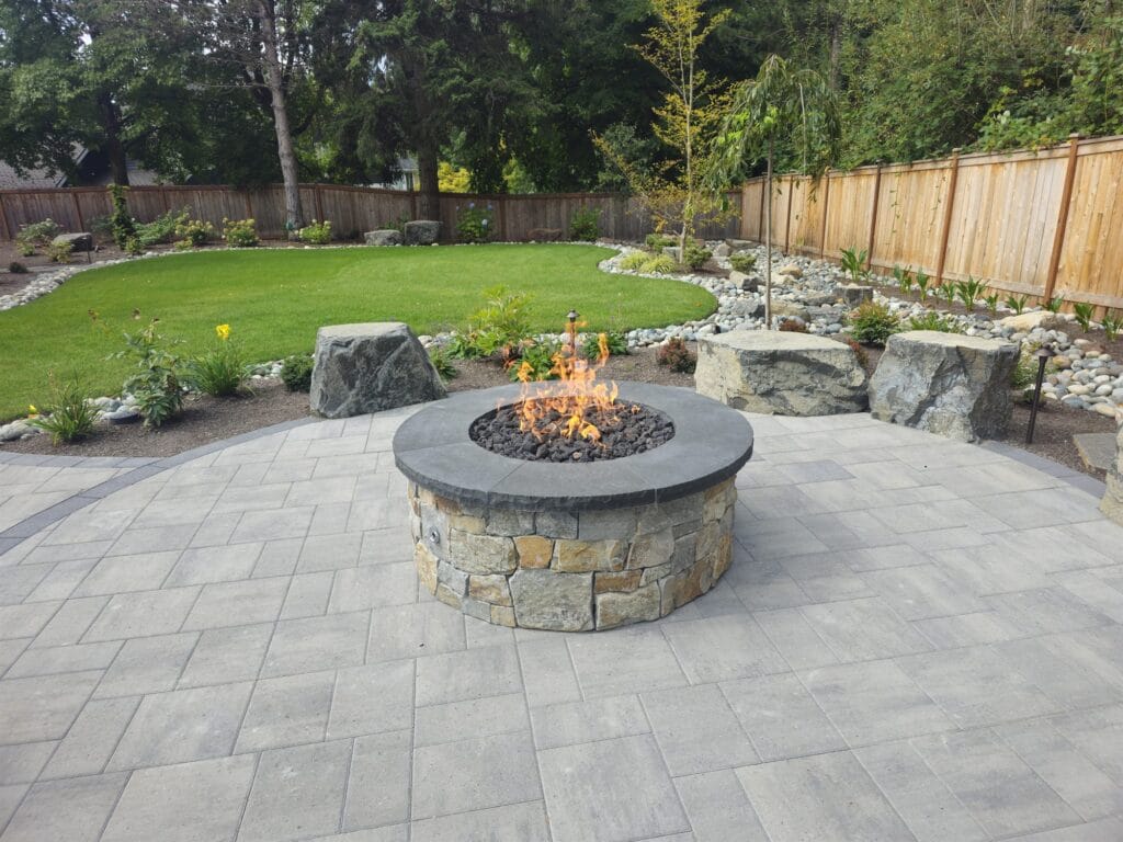 Round stacked-stone gas fire pit with a bluestone cap on a gray paver patio ringed by boulders and lawn