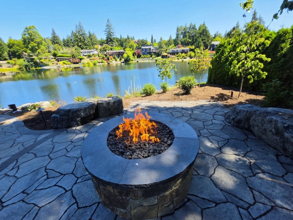 Round stone gas fire pit on a flagstone patio ringed by boulder seating, with a lake behind