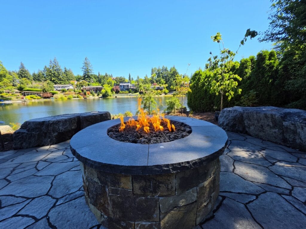 Round stone gas fire pit with a smooth stone cap on a flagstone patio, with boulder seating and a lake behind