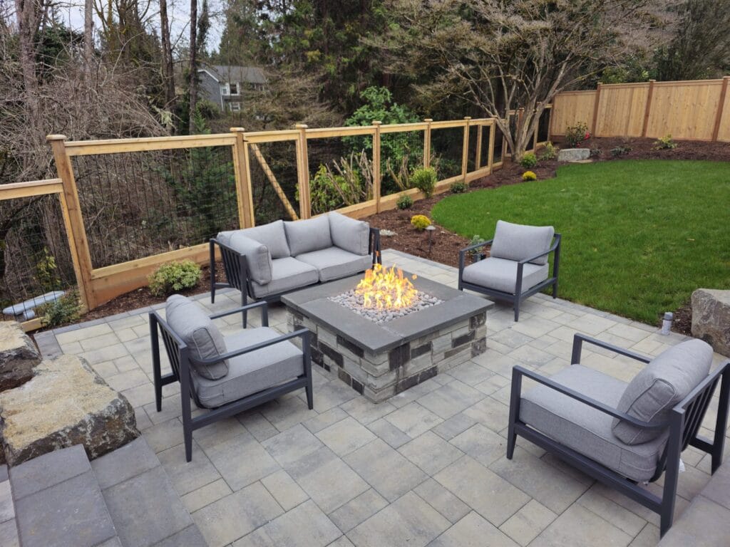 Square stone gas fire table with glass media surrounded by gray lounge chairs on a paver patio
